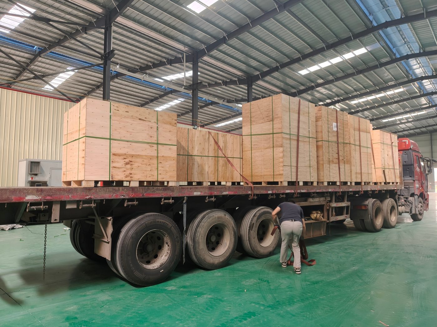 Plywood cases strapped to a flatbed trailer inside the loading bay, one operator tensioning the red lashing